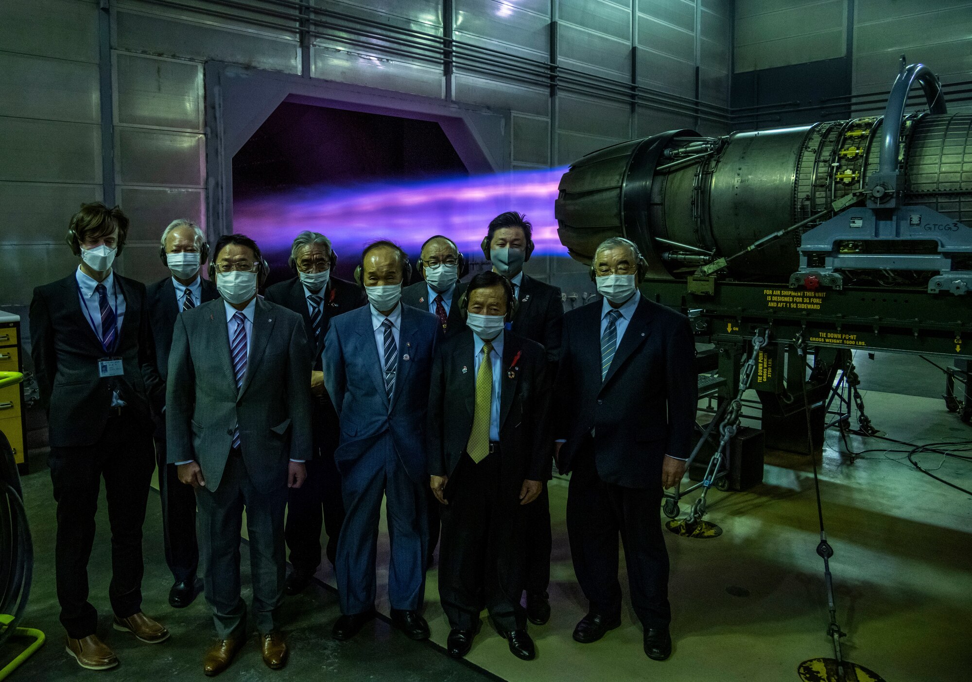 Men in suits stand in front of an F-16 engine.