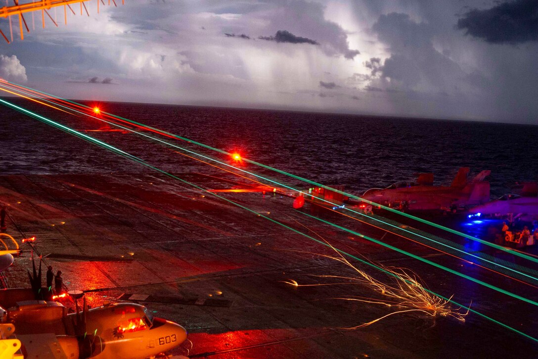 An aircraft lands on the flight deck of a ship at sea illuminated by colorful lights.
