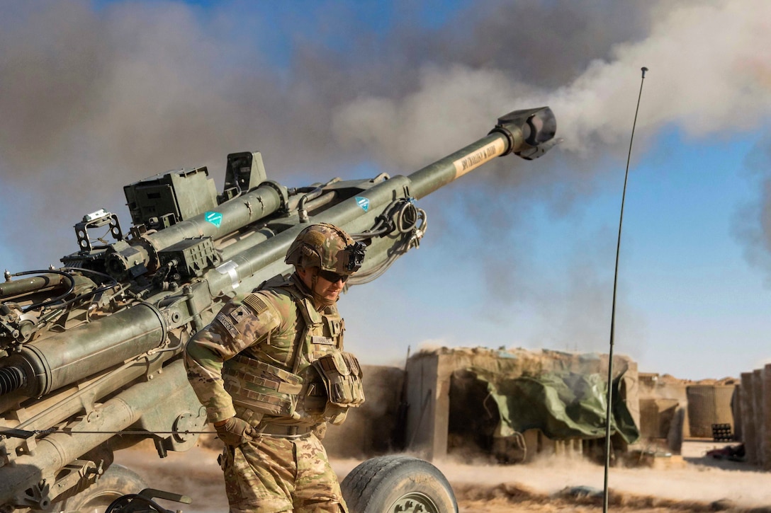 A soldier stands next to a weapon in a desert-like area surrounded by smoke.