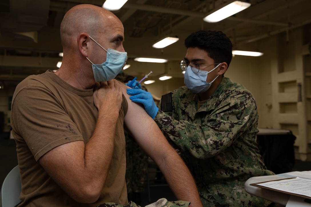 A sailor wearing a face mask and gloves holds a syringe and leans over to give a man wearing a face mask a COVID-19 vaccine.