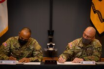 two men in army uniforms signing a memorandum on a table.