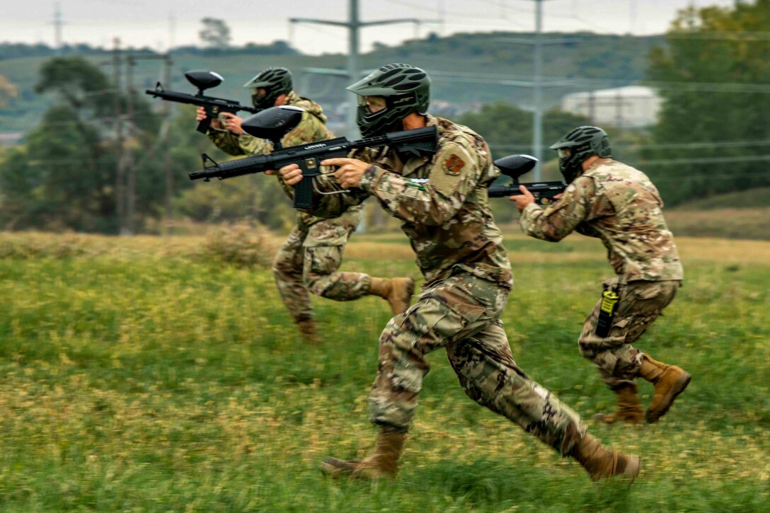 Three airmen run through a grassy field while holding weapons.