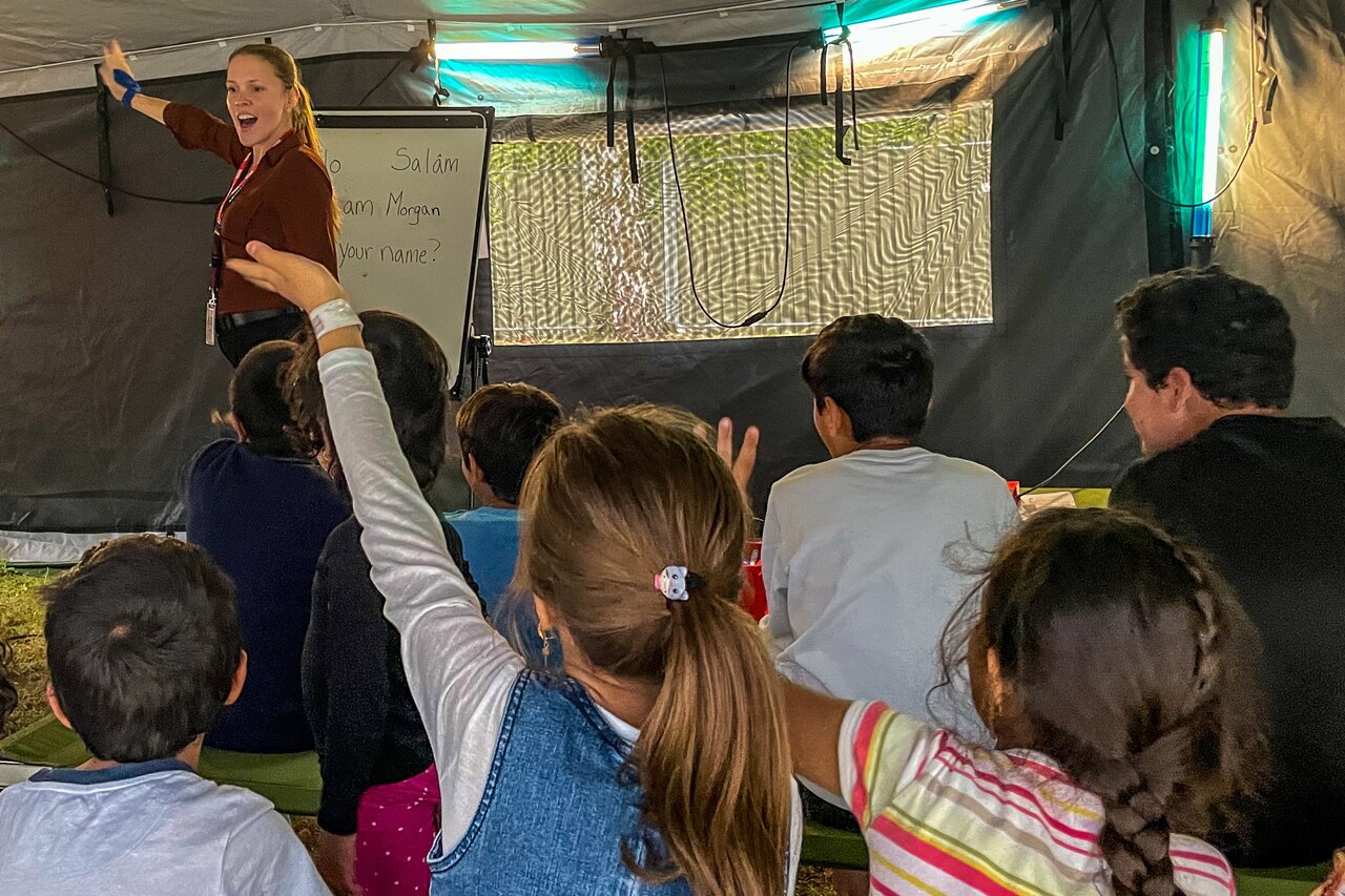Children sit and some raise their hands while a teacher stands in front of a room.