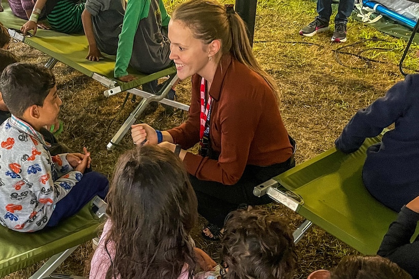 A teacher smiles while sitting on the ground with children.