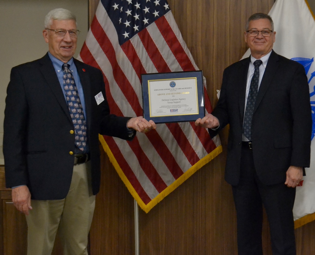 Two men in business suits hold a framed award between the two of them while standing in front of an American flag.