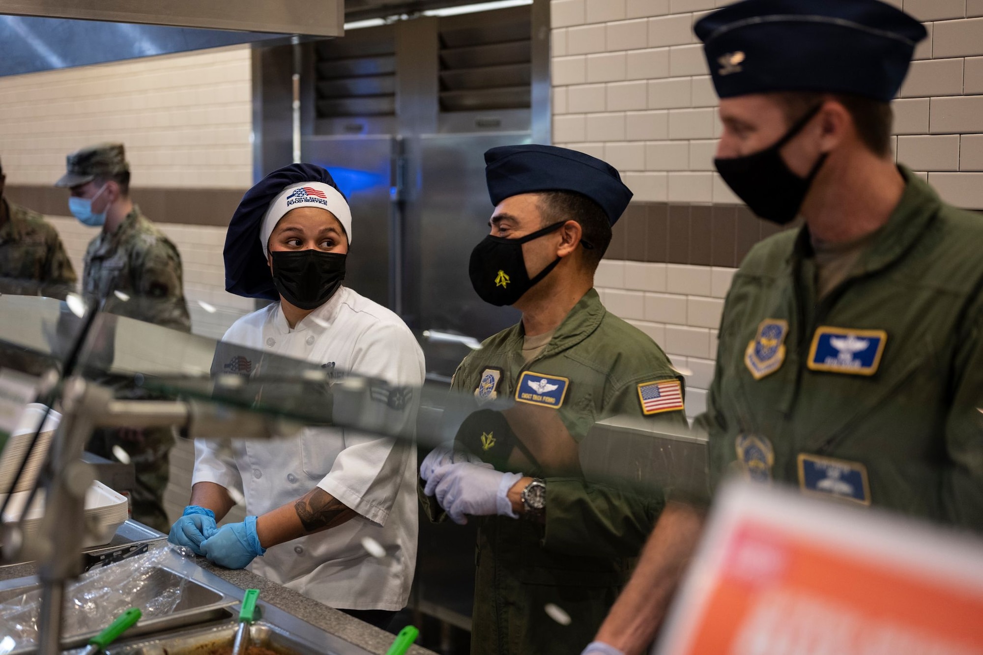 Two men in military uniforms talks to a woman in chef attire along a cafeteria-style line
