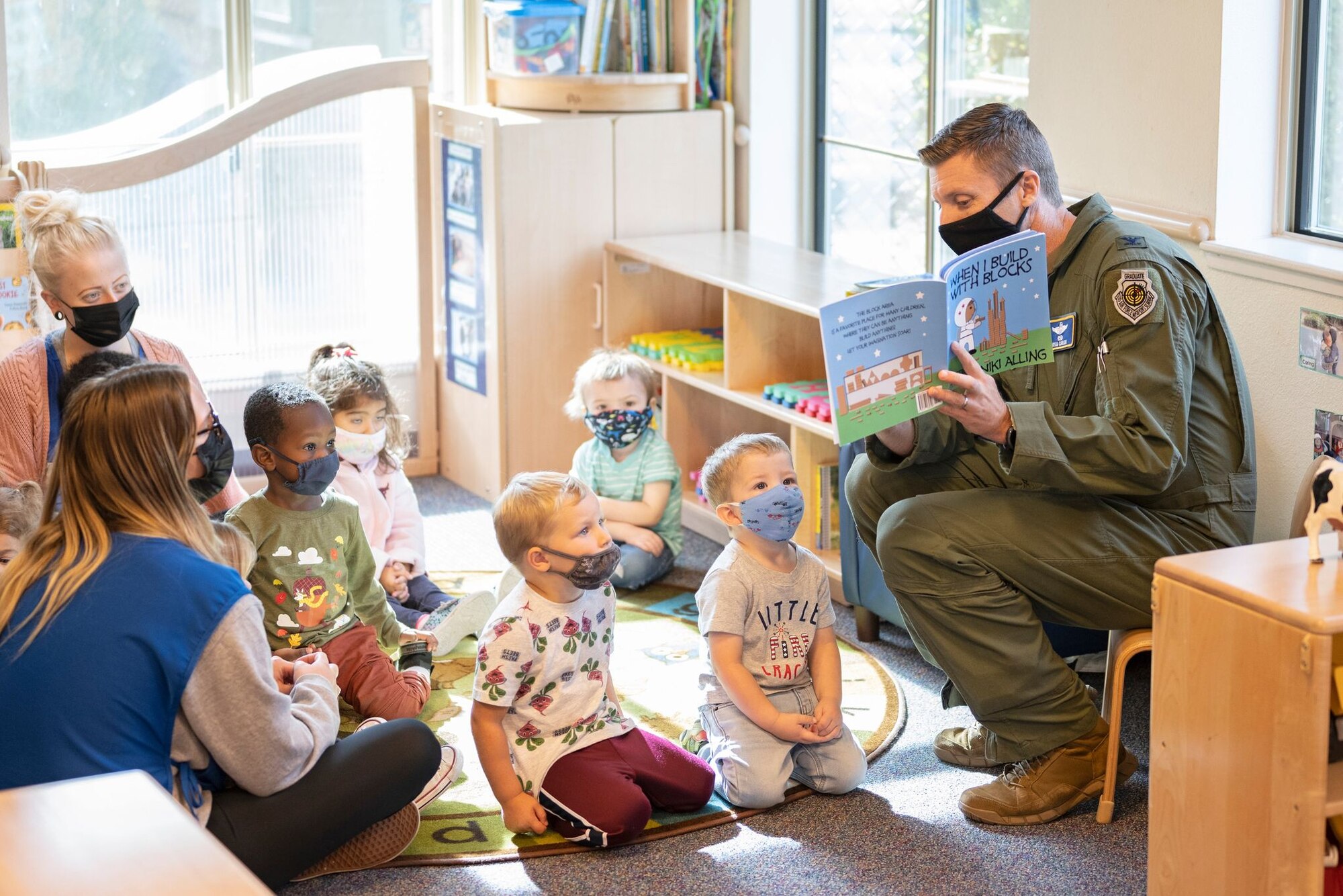A man in a military uniform reads a children's story to a gaggle of children in a brightly-lit classroom