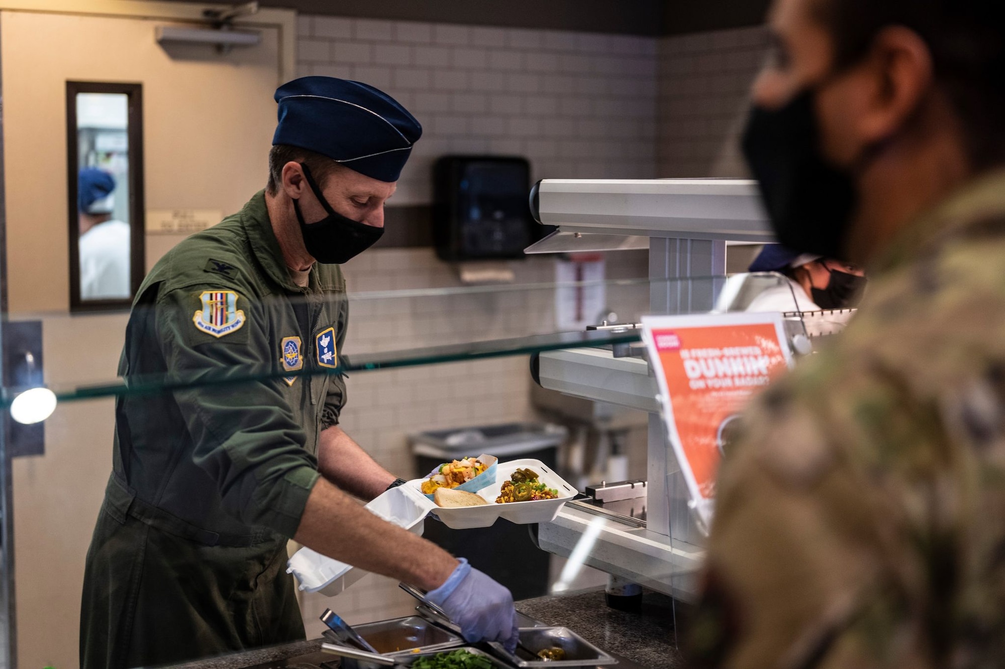 A man in a military uniform loads a to-go box with colorful food in a cafeteria-style line