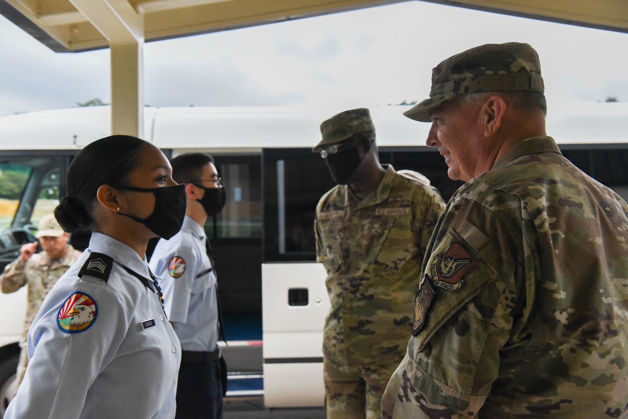 Two military members in uniform stand outside a school and talk to students in JROTC uniforms.