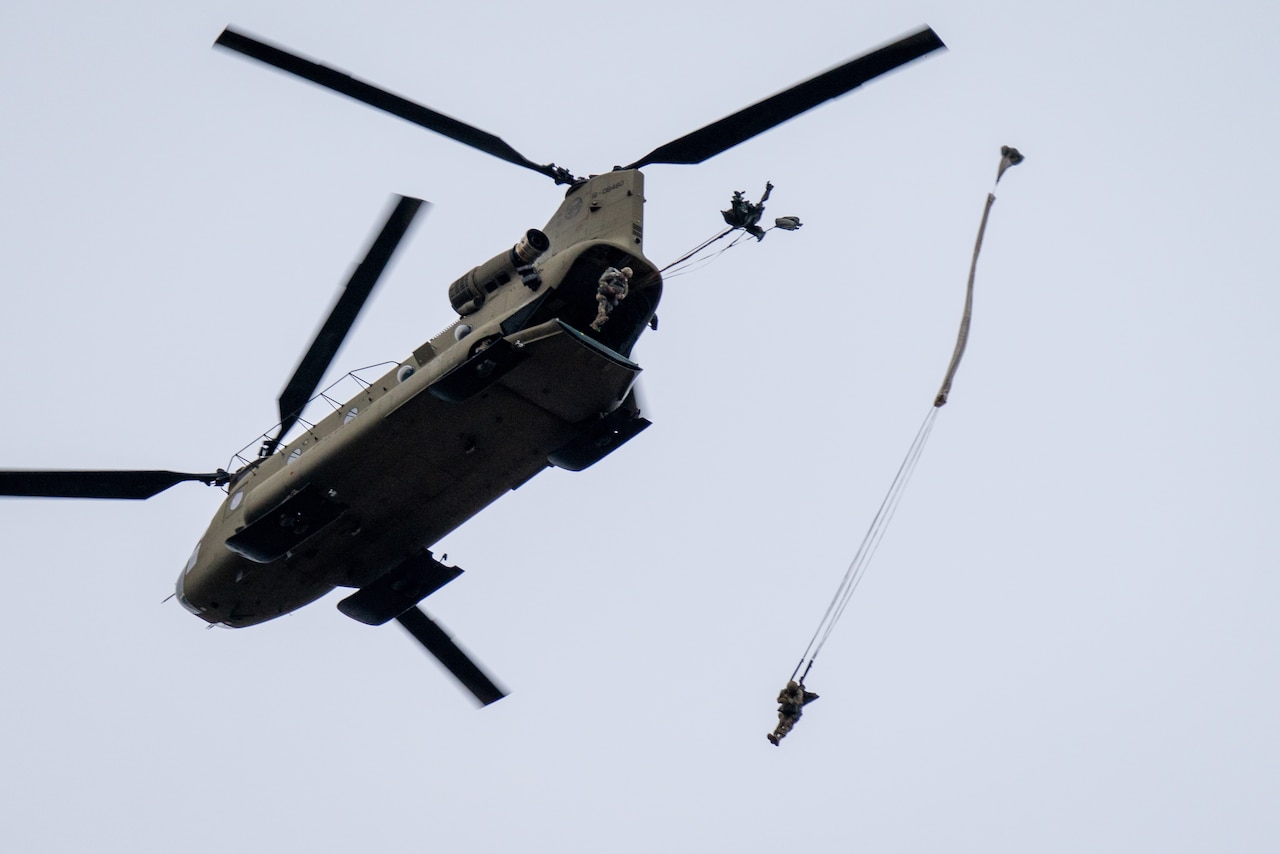 Two soldiers jump from an airborne helicopter while wearing parachutes.