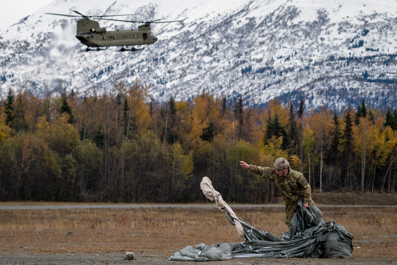 A soldier maneuvers a parachute as a helicopter flies in the background.