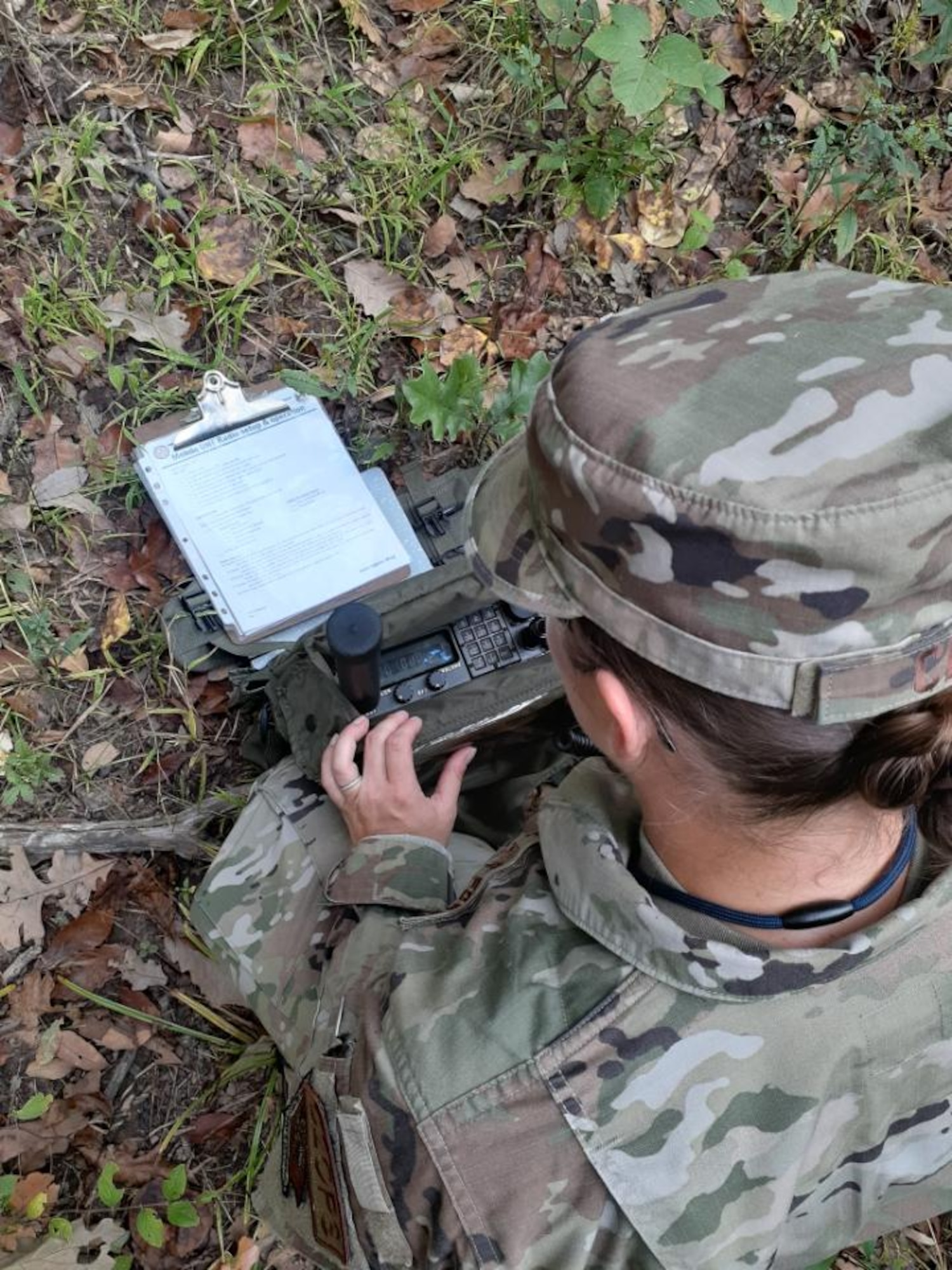 A woman in camouflage with her back to the camera kneels while holding a radio in front of her.