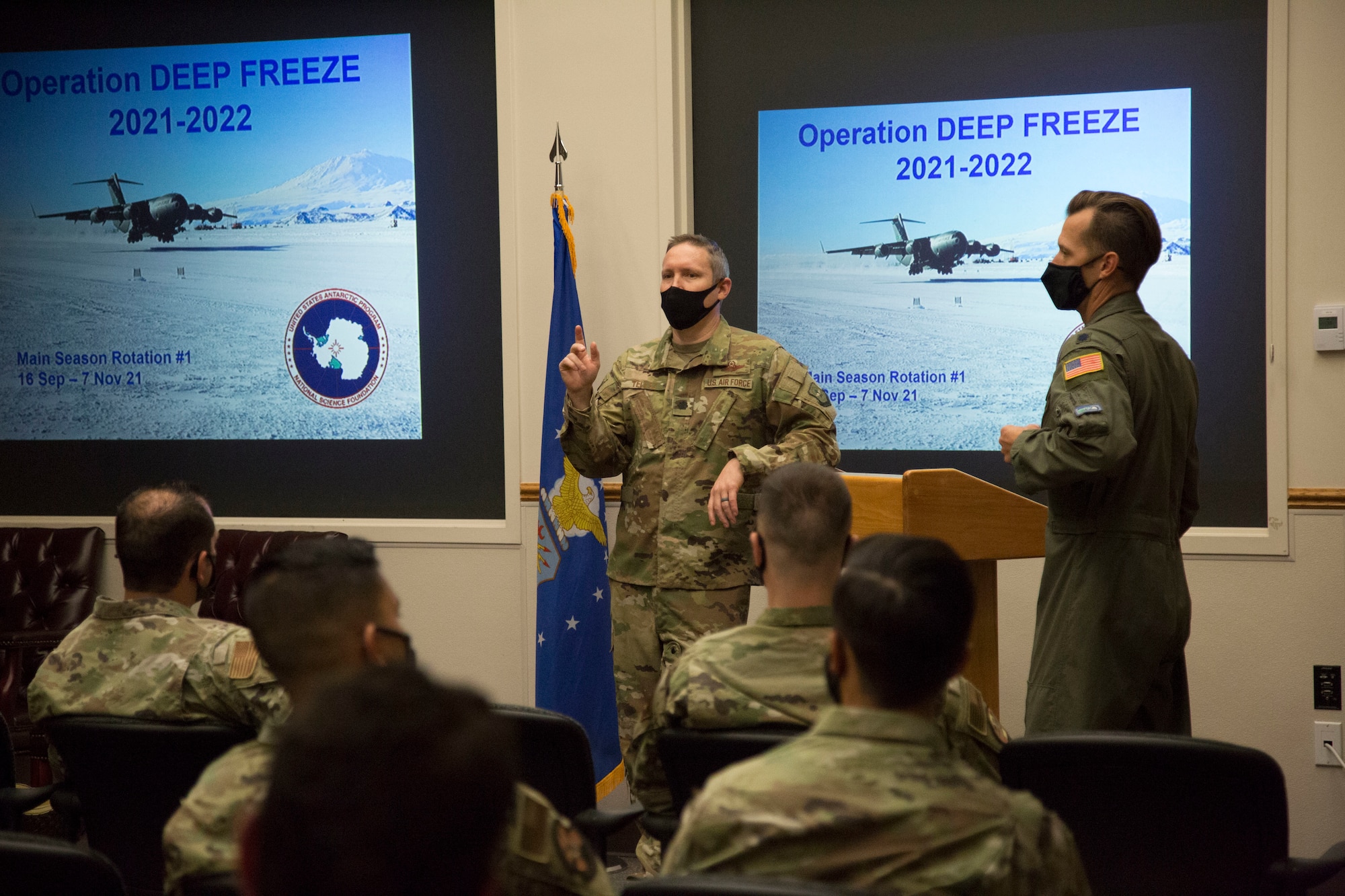 Two airmen at a podium briefing in front of a screen in an auditorium