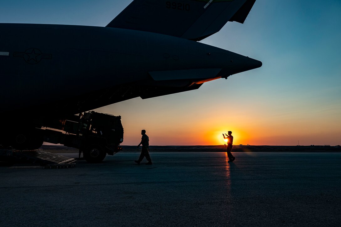 An airman guides a vehicle into an aircraft under a sunlit sky as shown in silhouette.