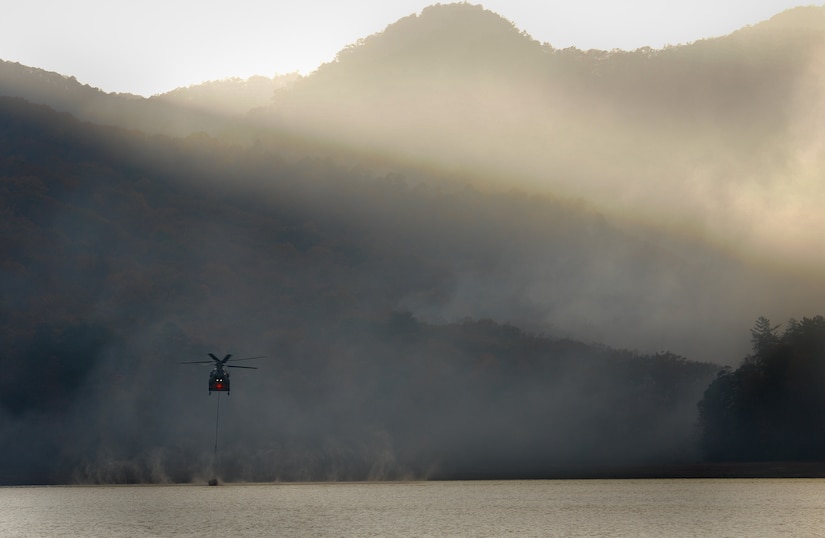 A helicopter scoops water from a lake, as smoke rises from behind.