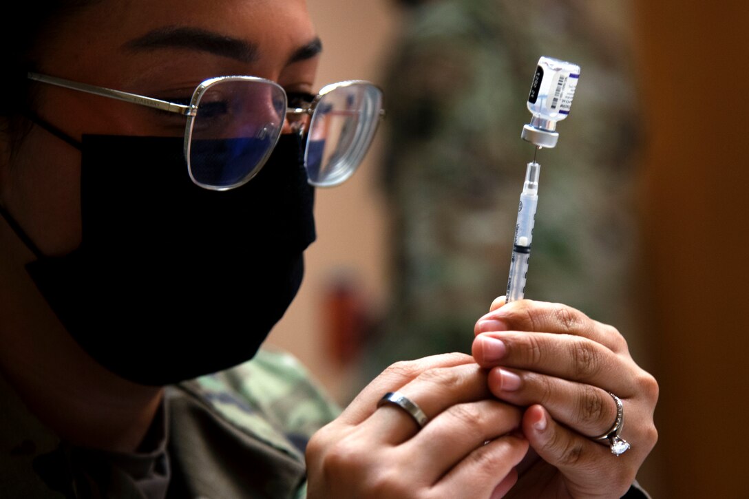 An airman wearing a face mask holds a syringe inserted into a small vaccine bottle.