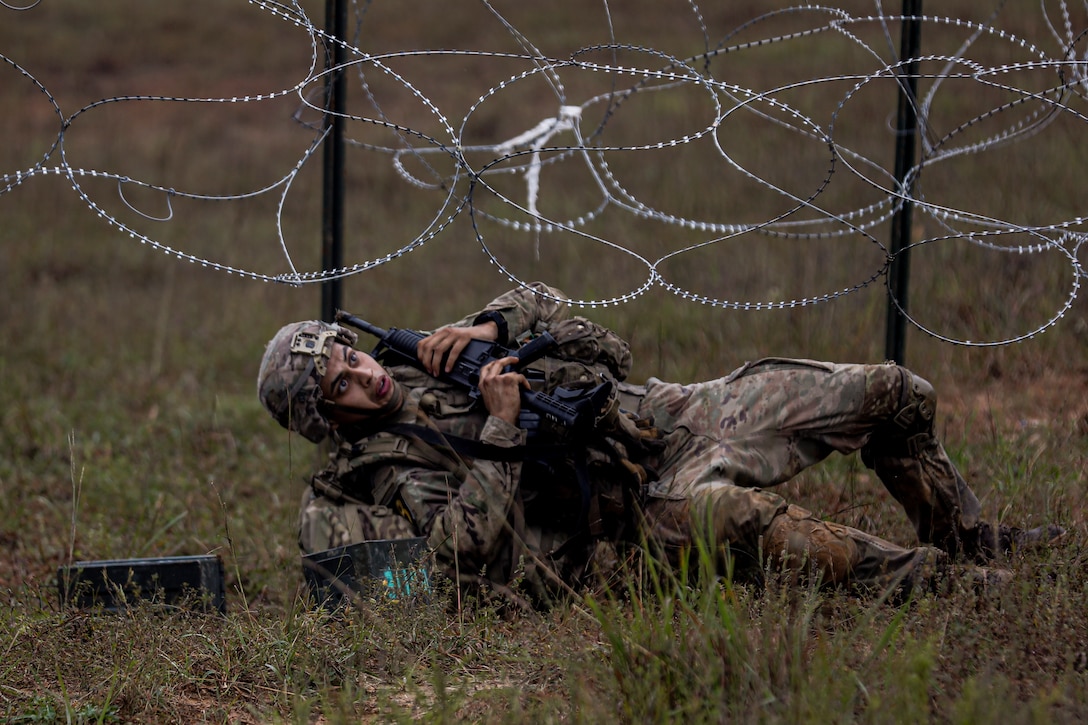 A soldier carrying a weapon lays on the ground and moves under razor wire.