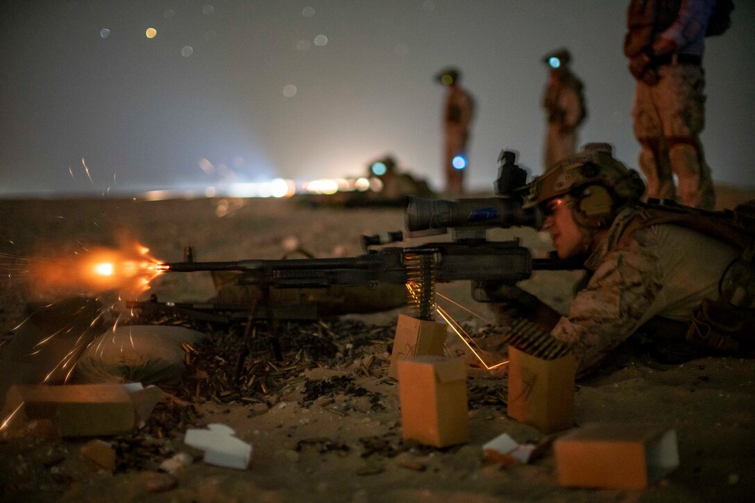 A Marine laying on the ground fires a weapon as fellow Marines observe under a starry sky.