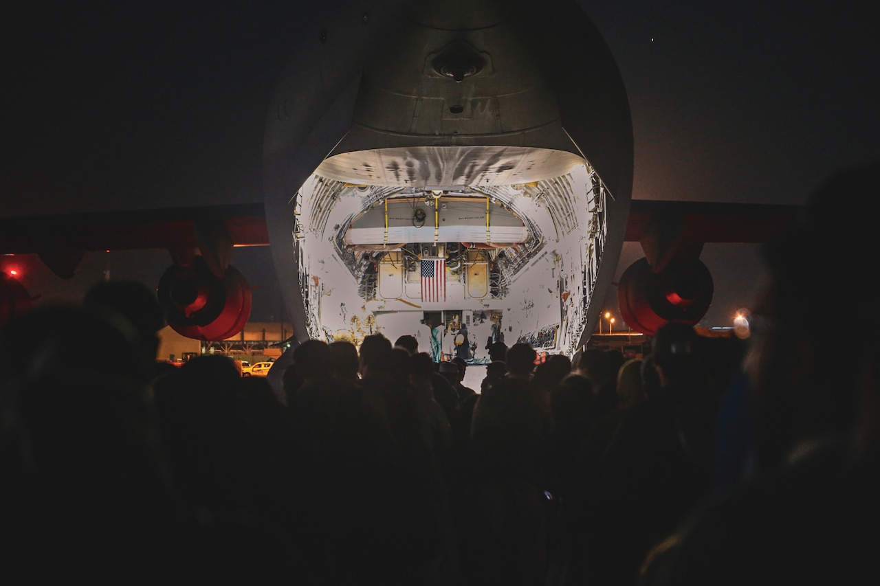 A crowd of people face the illuminated back of a C-17 at night.
