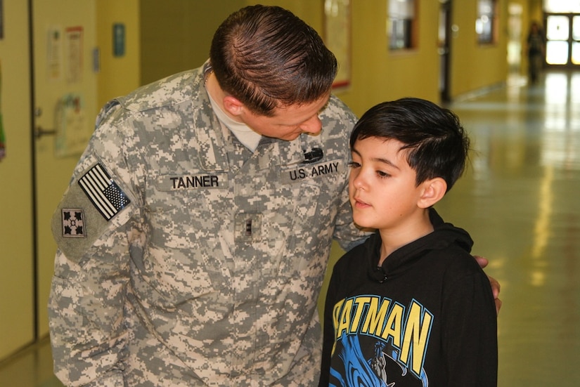 A soldier has his arm on the shoulder of a young boy as he leans forward to speak to the boy.