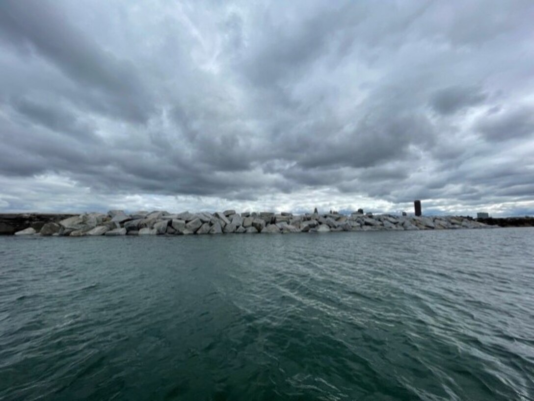 Large rocks piled to form a breakwater in water, with a city skyline in the background.