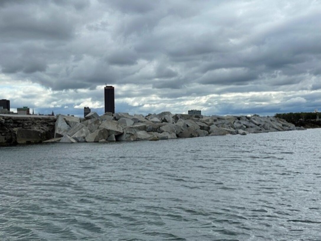 Large rocks piled to form a breakwater in water, with a city skyline in the background.