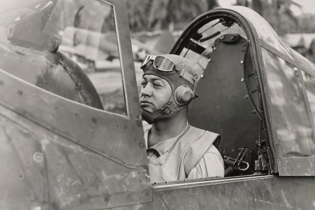 A man in aviator goggles and cap sits in an airplane cockpit.