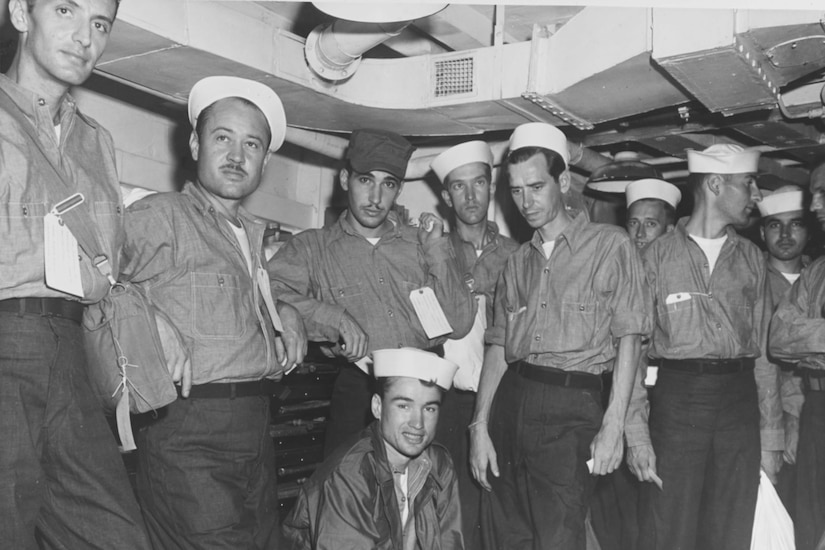Ten men in sailor uniforms stand close together in a compartment of a ship.