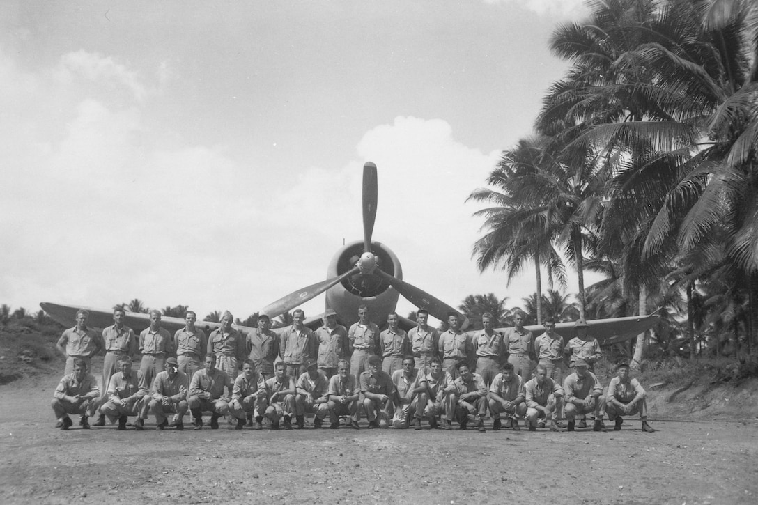 Two rows of men pose for a photo in front of a propeller plane on tarmac. Palm trees surround them.