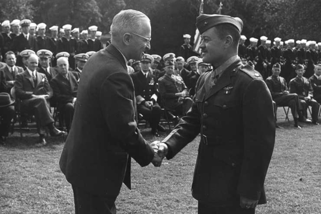 A man in a business suit and a man in a military uniform shake hands. An audience of civilian and military personnel looks on.