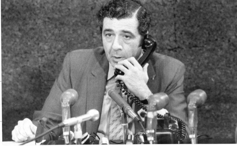 A man sitting in front of several microphones talks on a telephone.