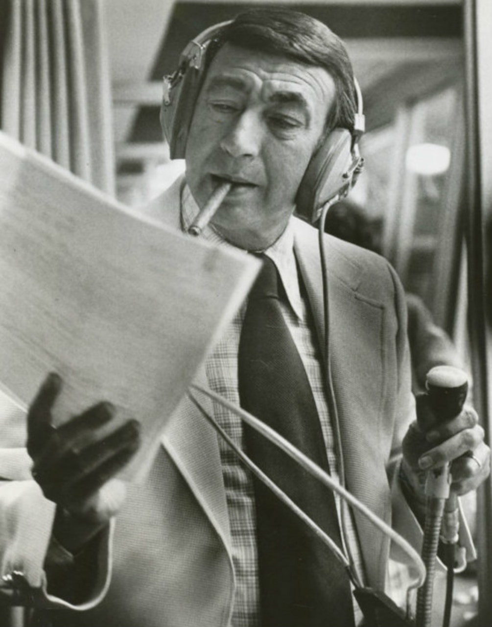 A sportscaster, wearing headphones and with a cigarette in his mouth, reads a script.