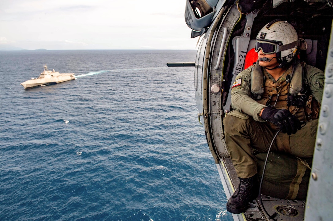 A sailor sits in a helicopter as it flies over an ocean near a ship.
