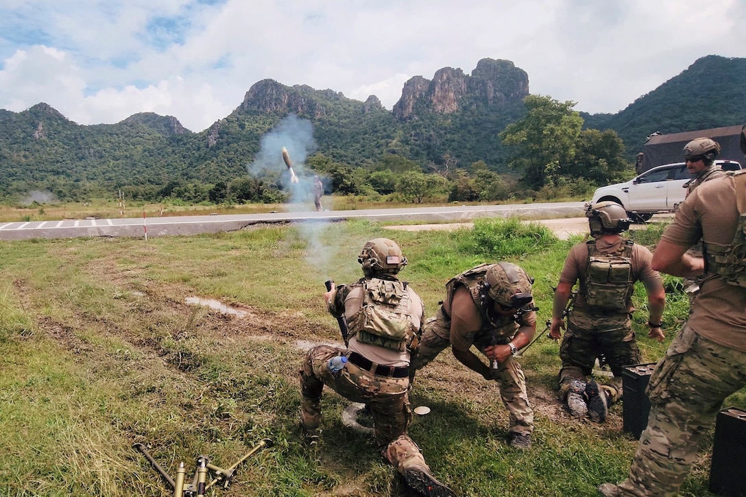 Soldiers fire a weapon in a field towards mountains.