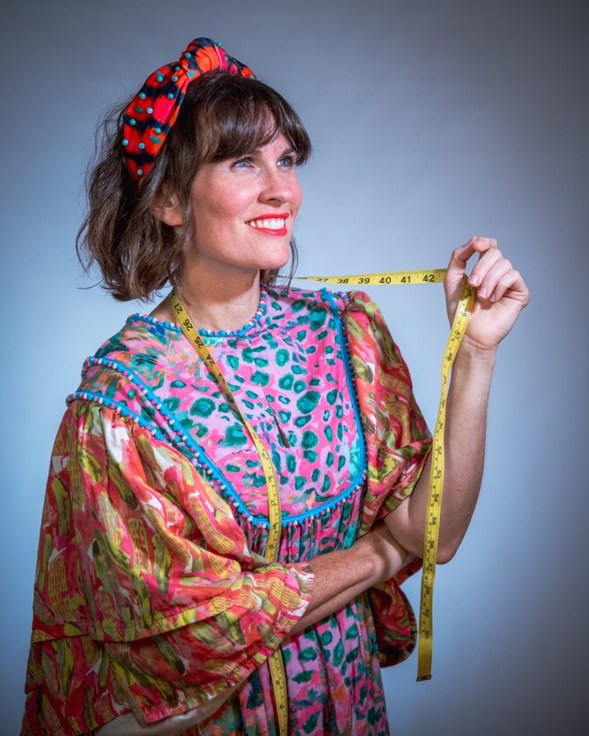 A woman dressed in colorful clothing looks into the distance while fingering a tape measure that's draped around her neck.