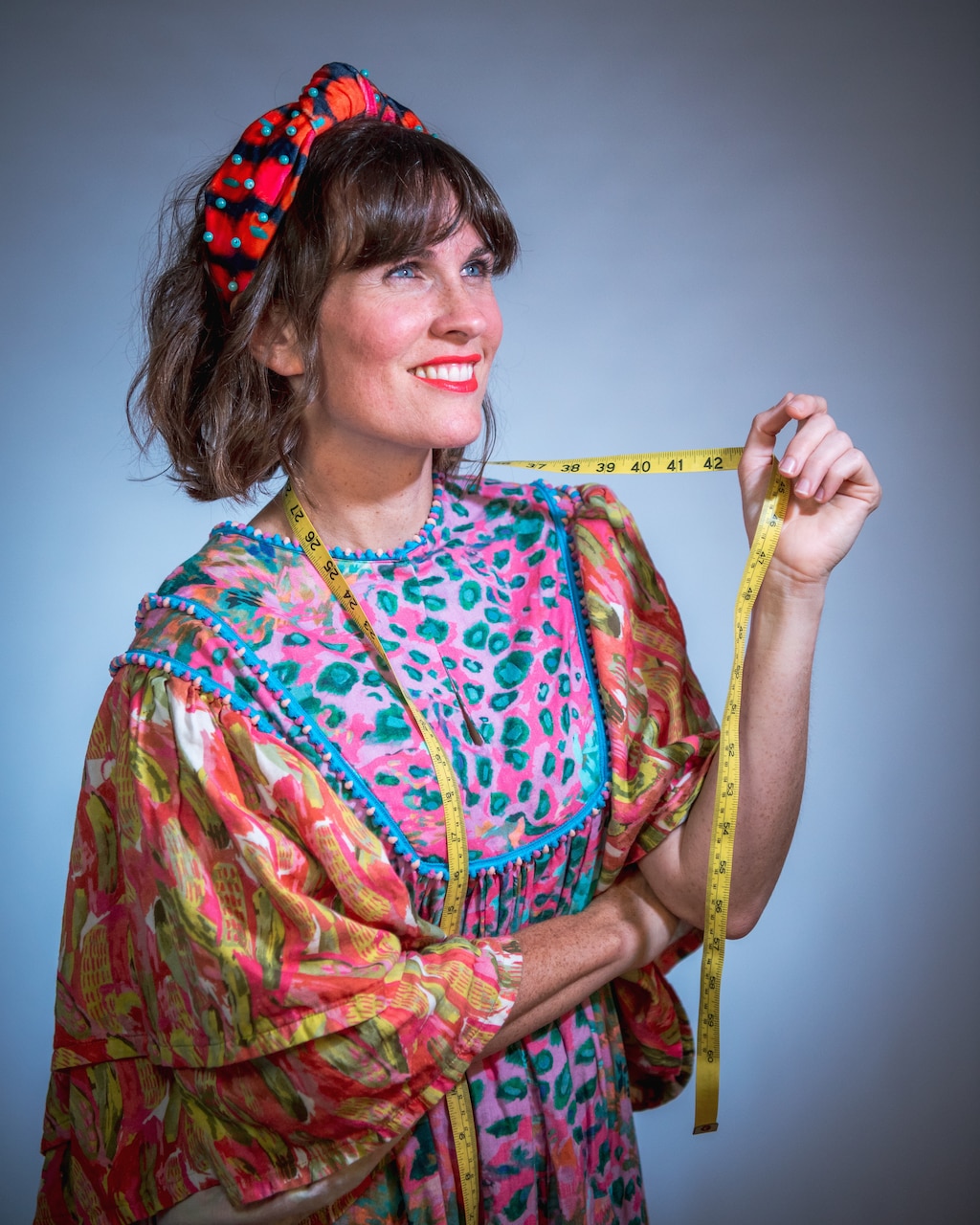 A woman dressed in colorful clothing looks into the distance while fingering a tape measure that's draped around her neck.