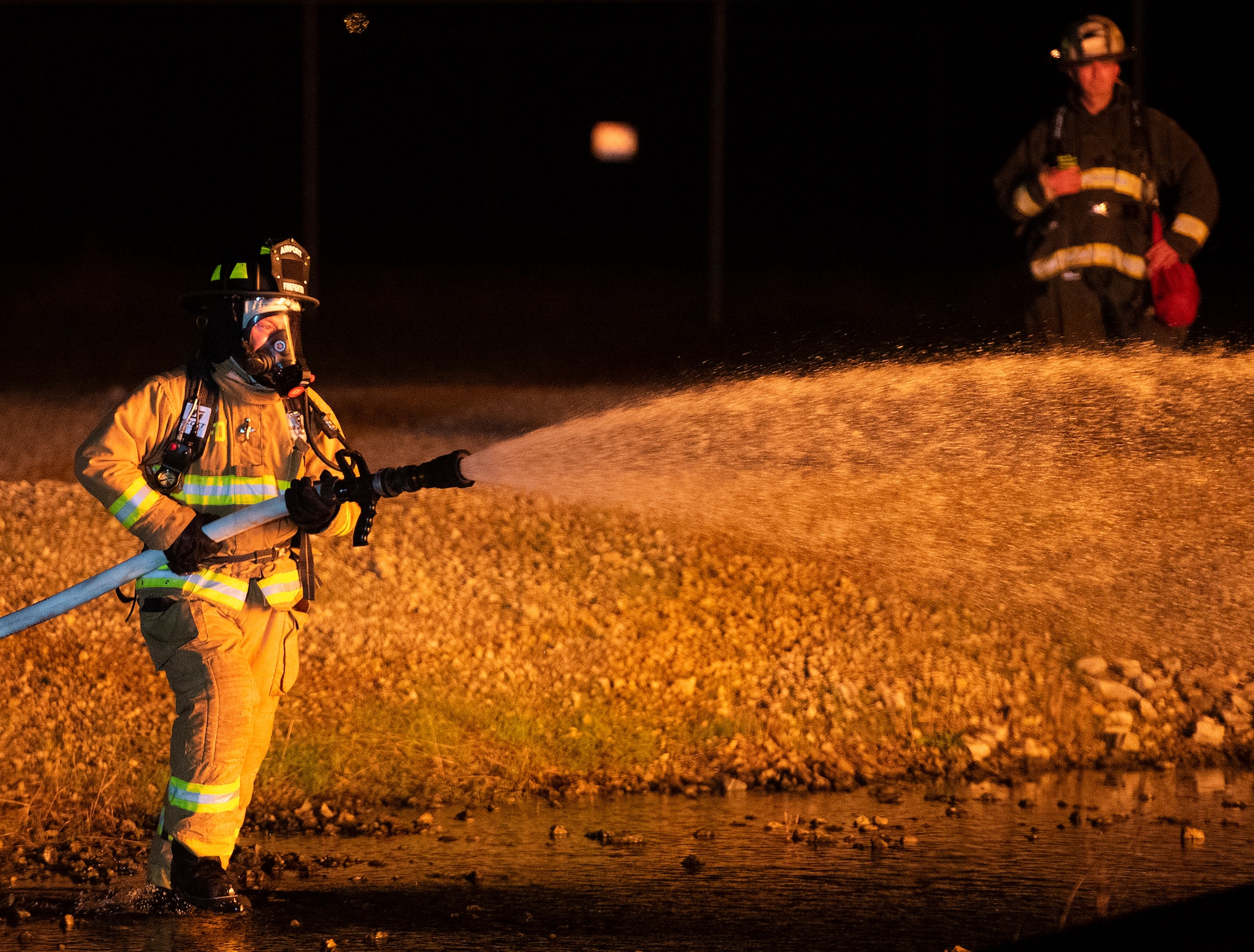 A Dayton International Airport firefighter operates a hose line against flames around an aircraft fuselage mock-up Oct. 5, 2021, at Wright-Patterson Air Force Base, Ohio. A 788th Civil Engineer Squadron firefighter monitors the training in the background. (U.S. Air Force photo by R.J. Oriez)
