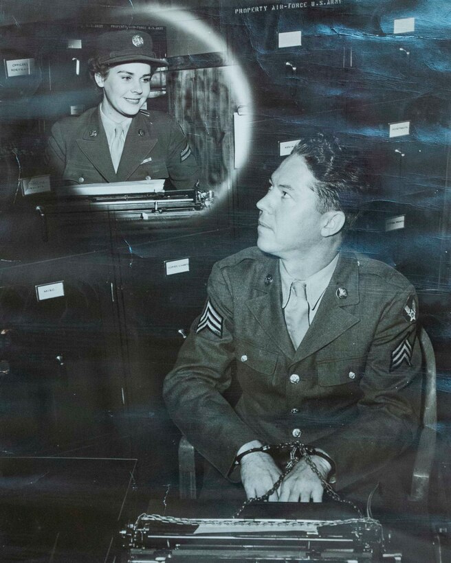 A World War II-era black-and-white recruitment photo shows a man at a typewriter looking up at a woman seated in front of a typewriter.