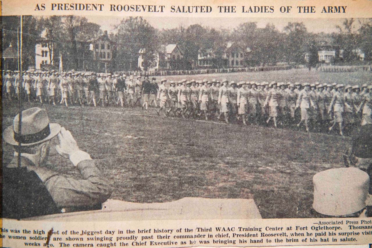 An old newspaper clipping of a man saluting women in military uniform is shown.