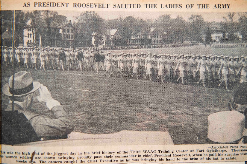 An old newspaper clipping of a man saluting women in military uniform is shown.