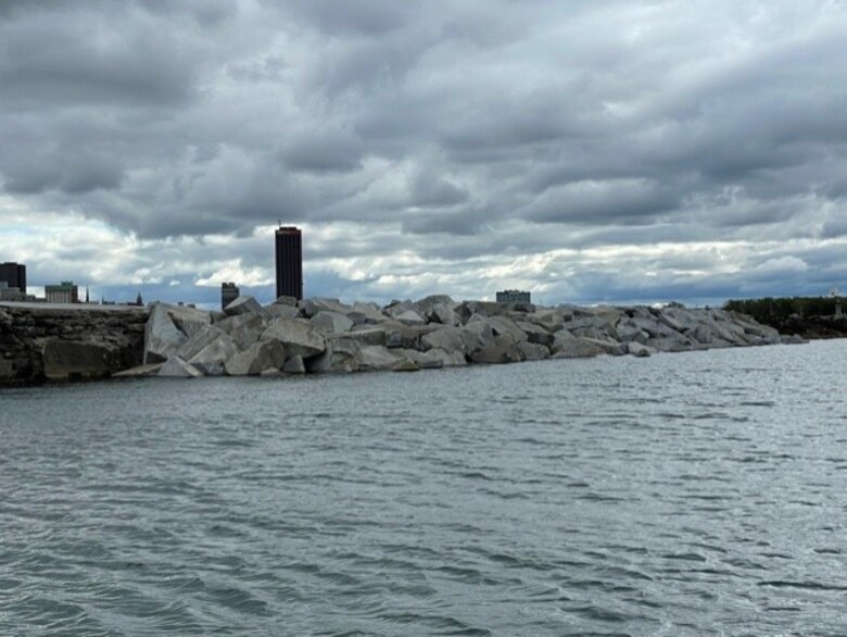Large rocks piled to form a breakwater in water, with a city skyline in the background.