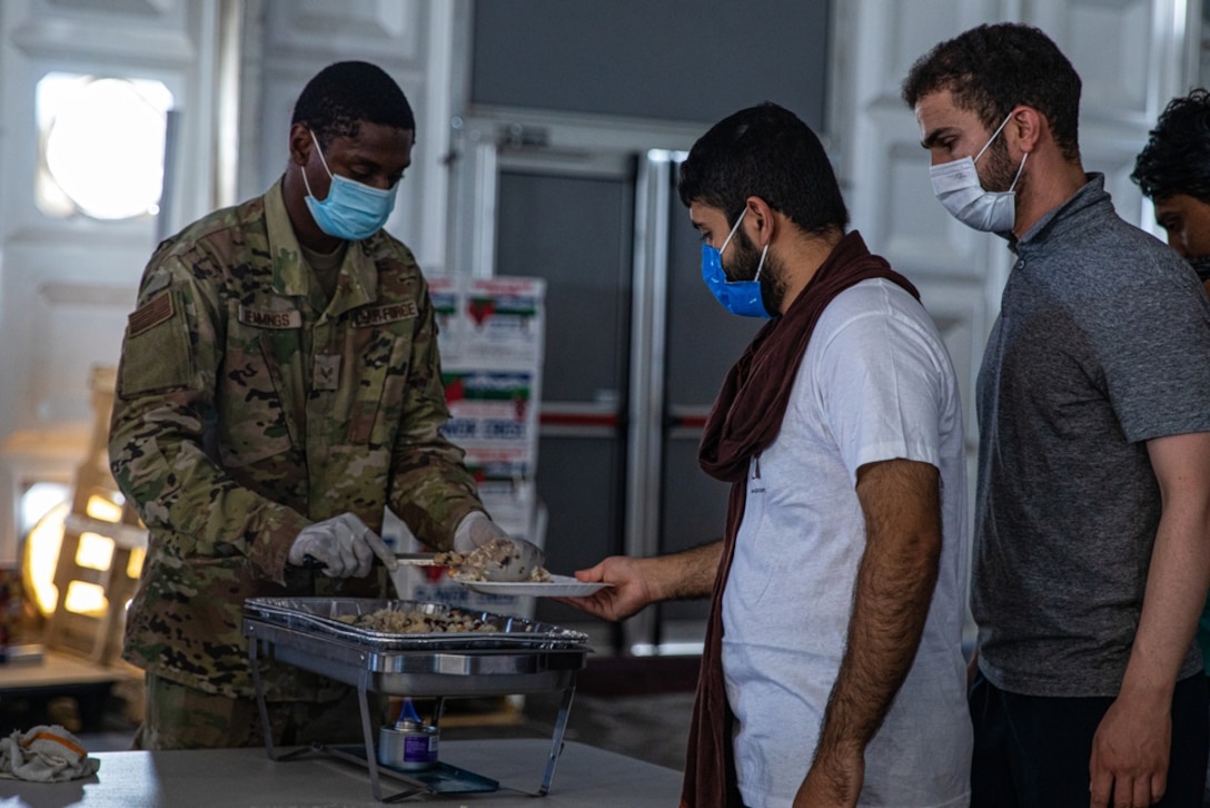 A military member in uniform serves food to two men lined up to receive a meal.