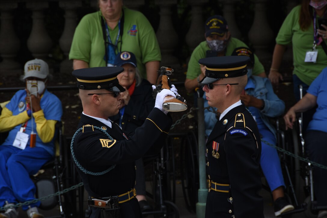 Four people sitting in wheelchairs watch two service members in dress uniforms.