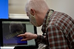 A man in a checkered shirt touches a computer screen to manipulate the 3D image of a spring.