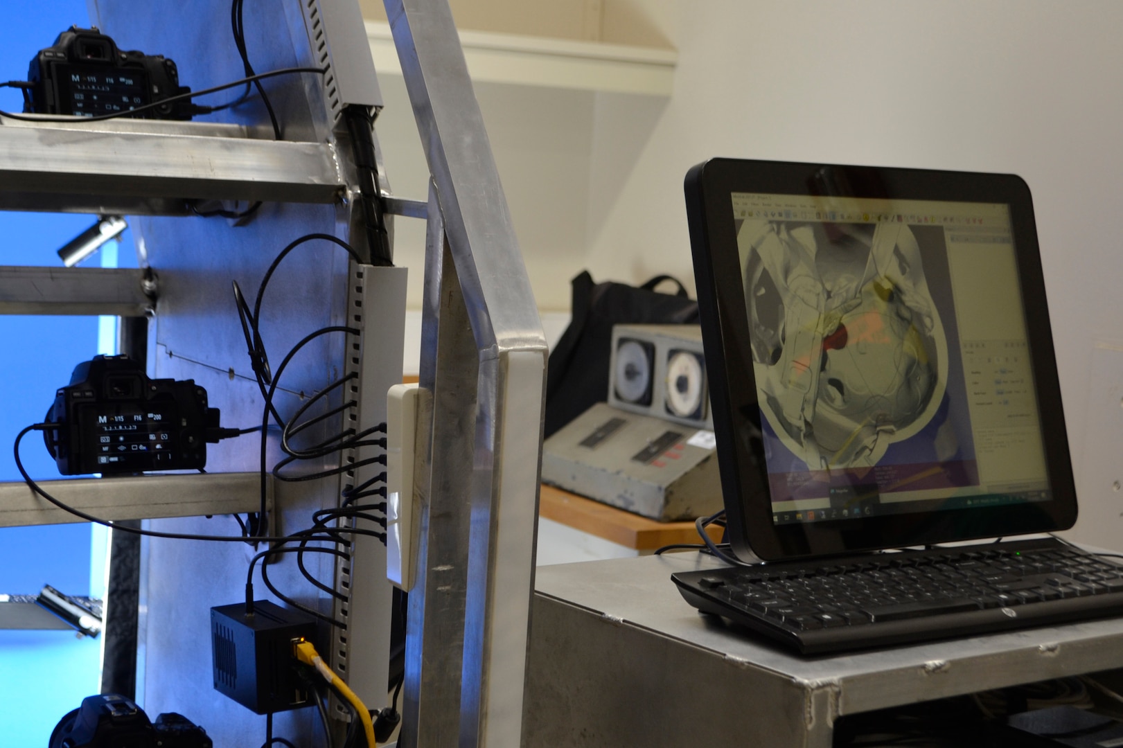 A computer screen on the right of the photo displays a 3D model of a military helmet. To the left, a large rack holds several cameras that make up a portion of a 3D scanning system.