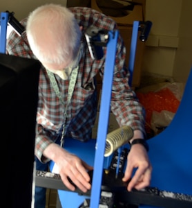 A man in a checkered shirt handles a rotating metal plate on which a coiled spring sits.