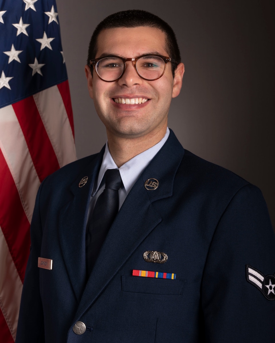 Official photo of A1C Cooper Johnson, horn with Heritage Winds and the Concert Band, two of six ensembles in the Air Force Heritage of America Band, Langley AFB, VA.  A1C Johnson is wearing blue service dress in front of an American flag.