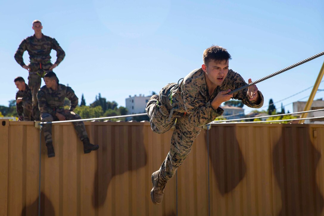 A Marine pulls himself along a rope while other Marines observe.