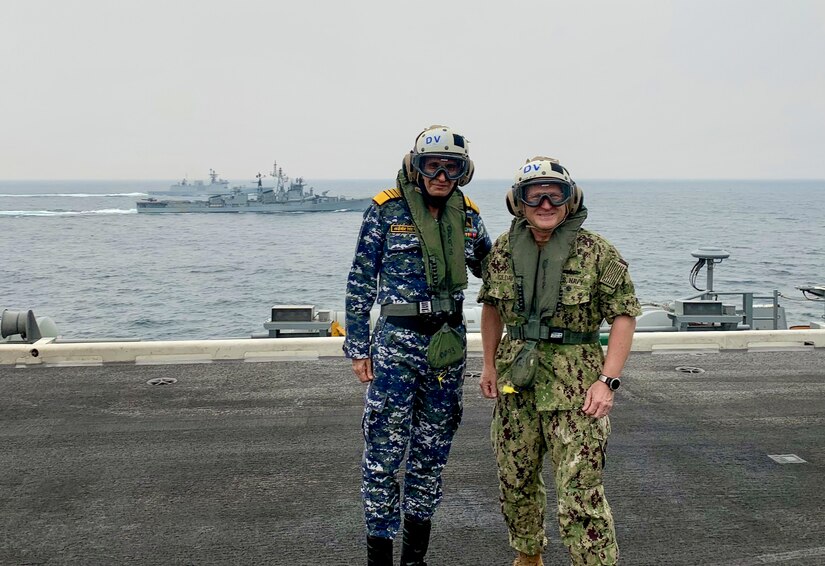 Two men in military uniforms stand on the flight deck of an aircraft carrier.