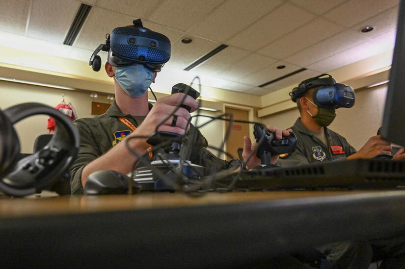Two men sit at computers wearing virtual reality goggles.
